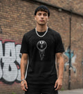 A young man with short dark hair and a black beanie stands outdoors against a graffiti-covered brick wall. He is wearing a black T-shirt featuring a detailed, circular graphic of a large black 8-ball with a small figure parachuting from it underneath. He wears a heavy silver chain and bracelet, enhancing the gritty, urban skater aesthetic.