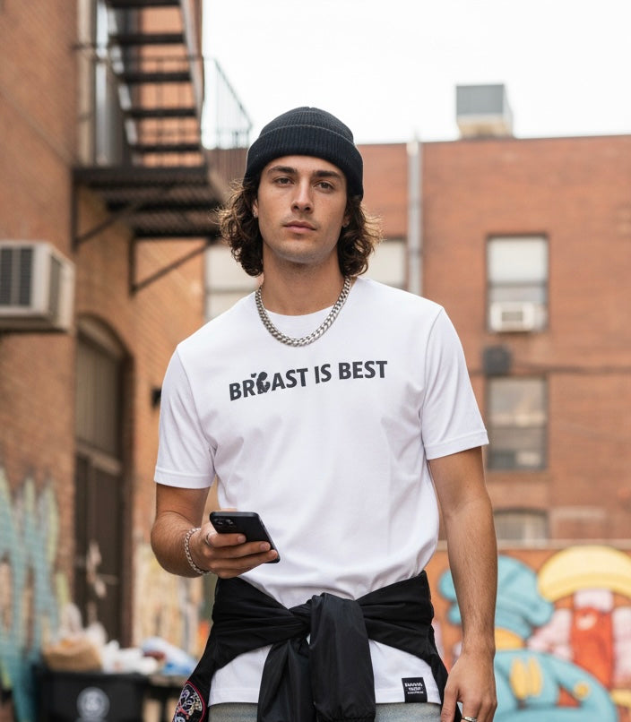 A young man with long wavy hair and a black beanie stands in a gritty urban alleyway, holding a phone. He is wearing a white T-shirt that boldly reads "BREAST IS BEST" in black text, with a small heart icon substituting for the 'E' in 'BREAST.' He has a silver chain and a jacket tied around his waist. The setting includes brick buildings and graffiti, embodying a cool, anti-establishment, skater-meets-hippie aesthe